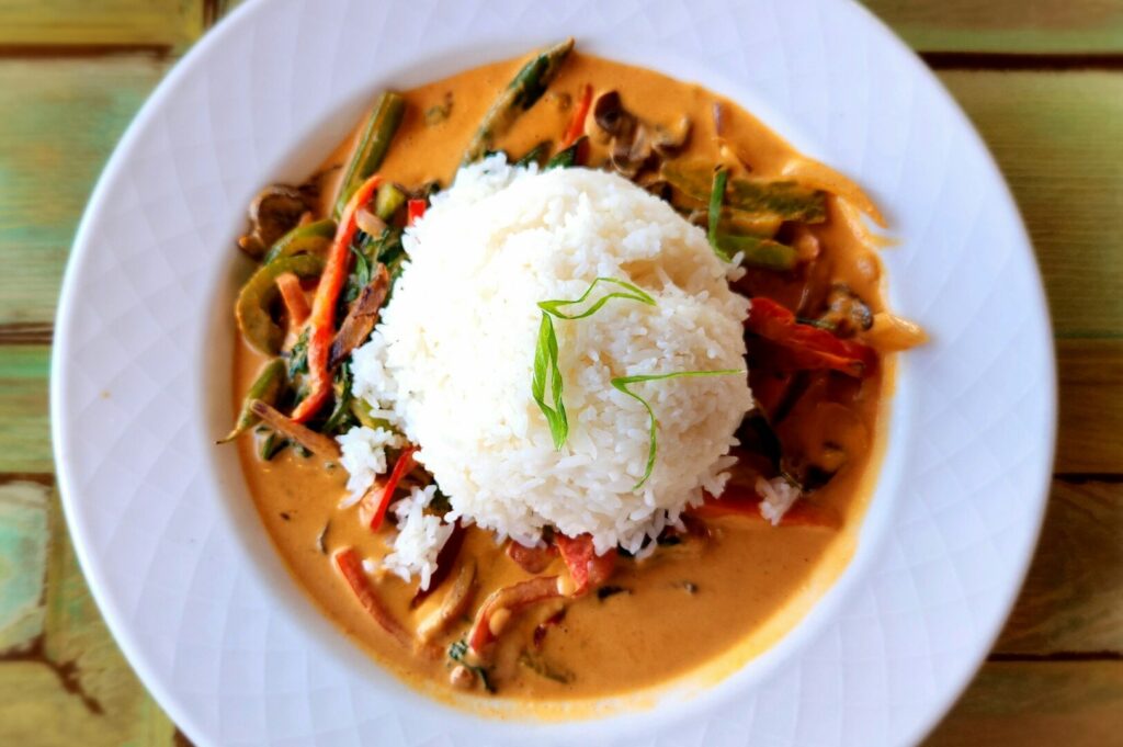 A white plate featuring a serving of rice topped with vibrant vegetables, representing a Thai curry meal.