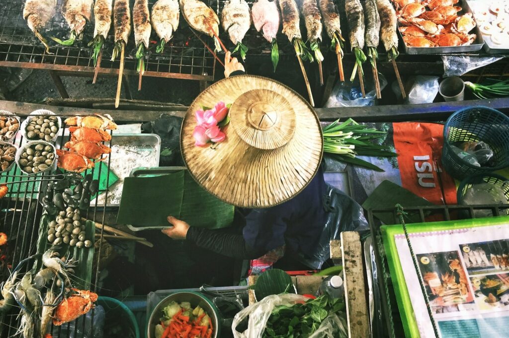 A woman in a hat stands in front of a vibrant market filled with various food items during a Bangkok food tour.