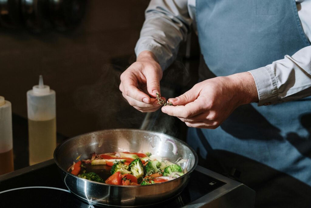 A chef wearing a blue apron carefully garnishes a steaming skillet of sautéed vegetables by sprinkling a sprig of fresh herbs into the mix. The stainless steel pan holds a vibrant combination of broccoli, red peppers, and onions, capturing a moment of precise culinary preparation in a professional kitchen.