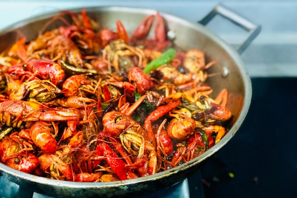 A large stainless steel pan is filled to the brim with bright red cooked crawfish, intermingled with dark spices and green chili peppers. The vibrant seafood dish sits on a stovetop, suggesting a spicy and flavorful preparation style like a traditional boil or Sichuan stir-fry.