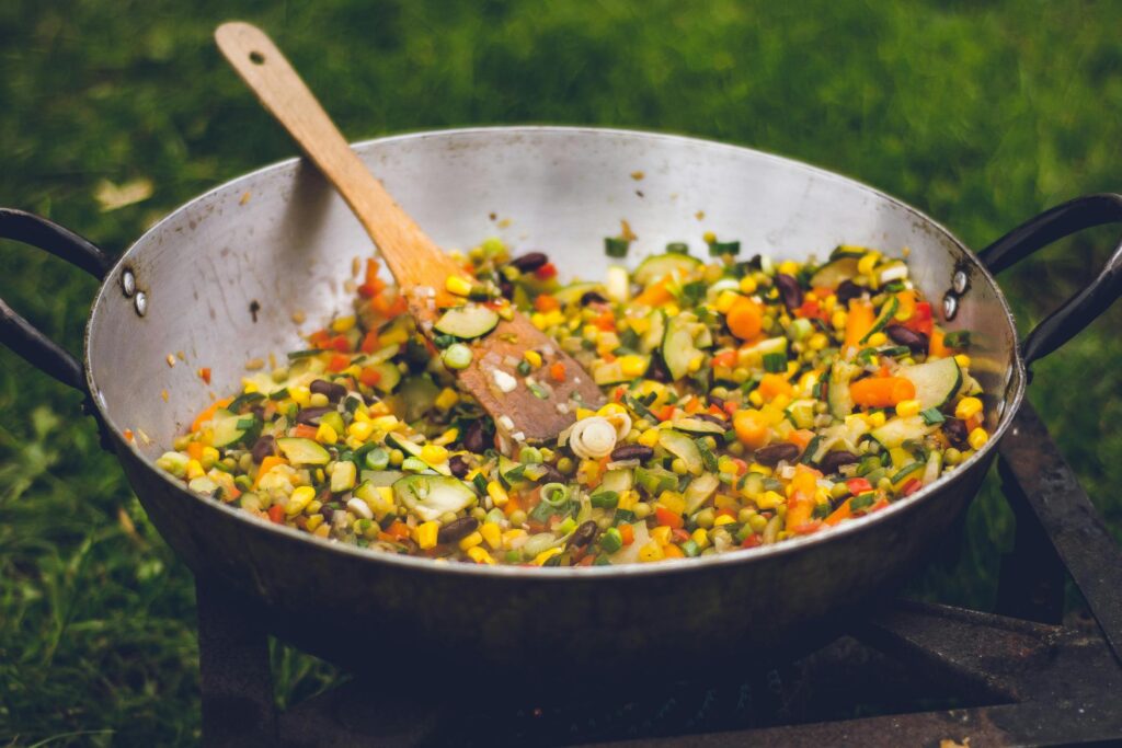 A large metal pan rests on a stand outdoors over green grass, filled with a colorful sauté of corn, zucchini, carrots, and red kidney beans. A wooden spoon lies amidst the vibrant vegetable mixture, suggesting a rustic, open-air cooking session.