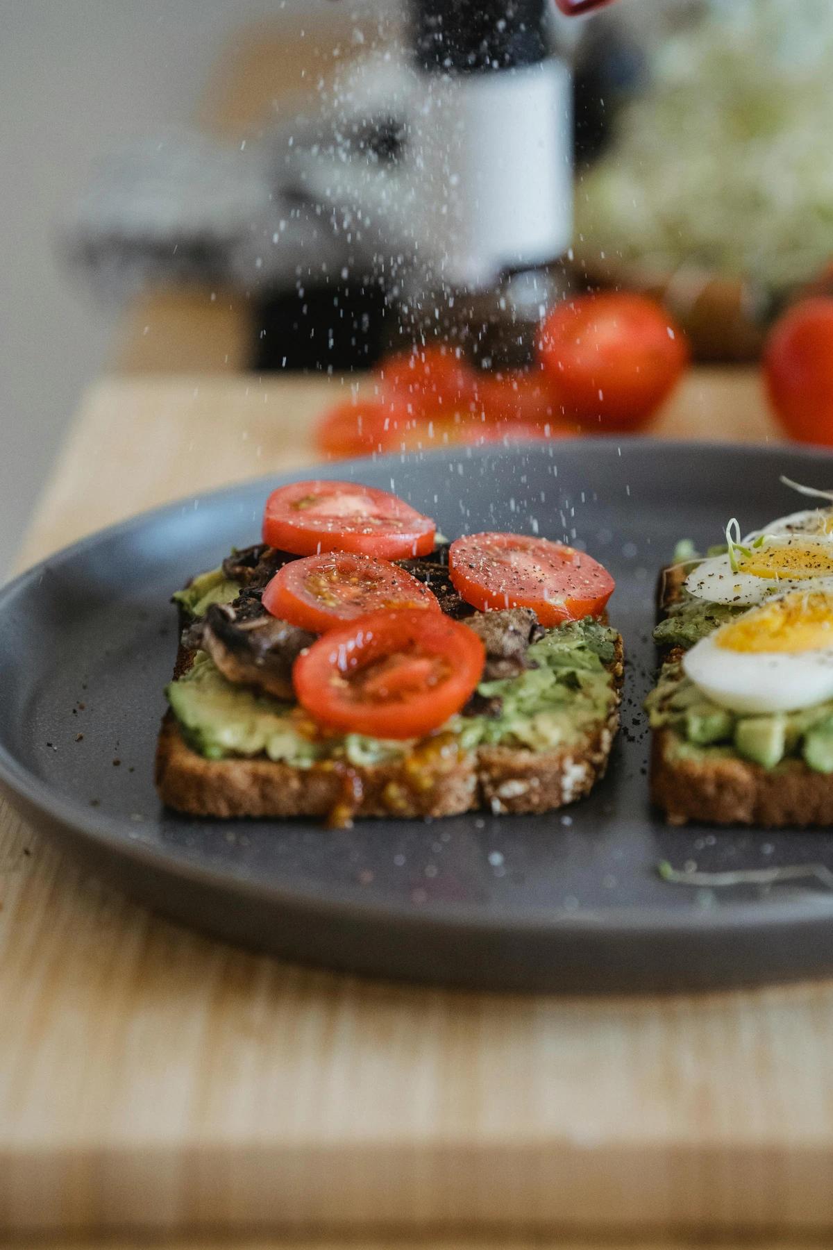 Close-up of avocado toast with tomatoes and eggs being seasoned, showcasing fresh ingredients and simple breakfast recipes.