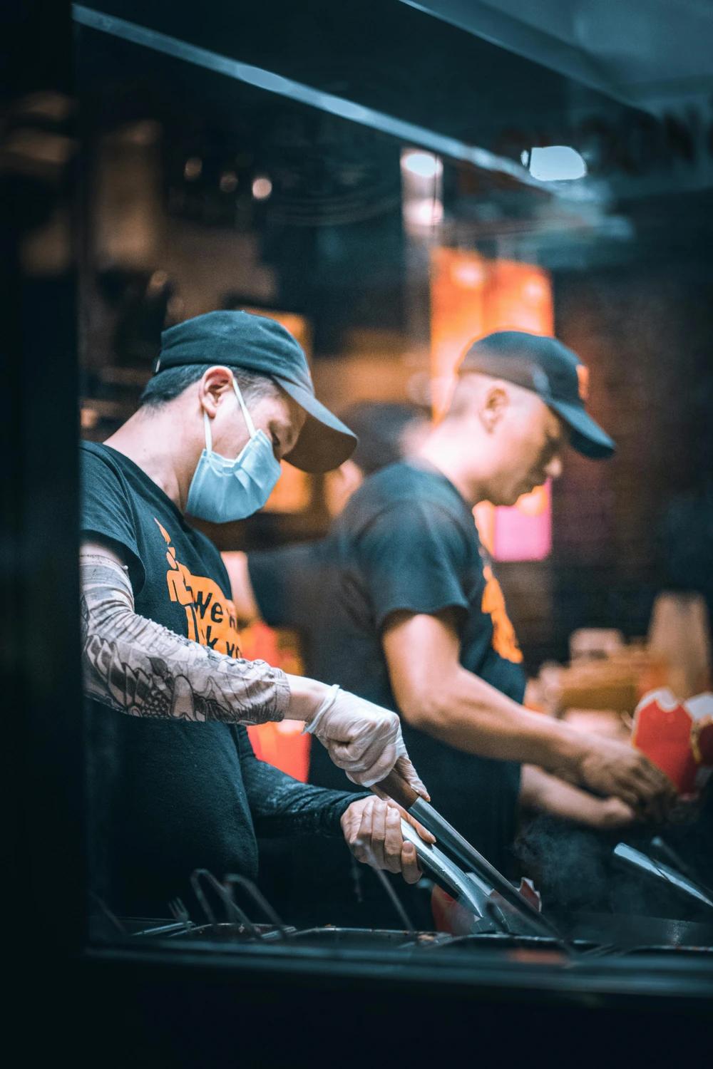 Two chefs wearing black t-shirts, baseball caps, and face masks working in a dimly lit professional kitchen, with one focused on using tongs at a cooking station.