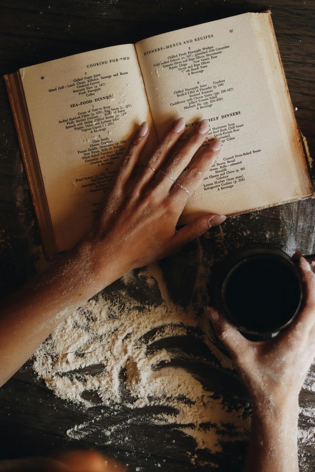Hands flipping through a vintage recipe book on a flour-dusted table, symbolizing home cooking inspiration and traditional recipes.