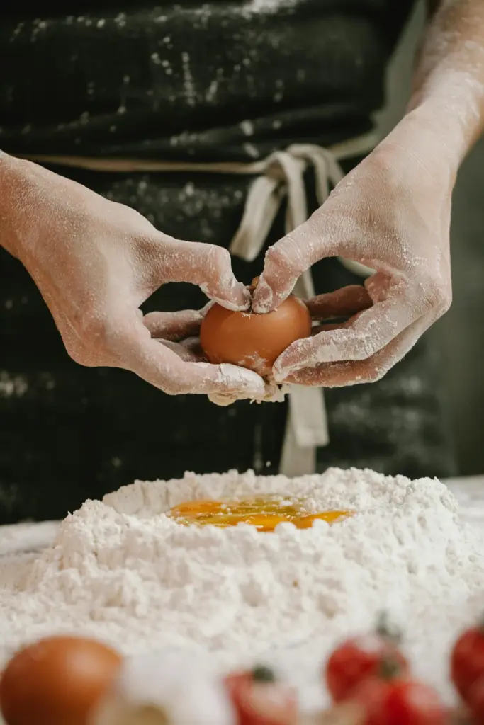 A close-up shot of a person's flour-dusted hands cracking a brown egg into a well of white flour on a wooden surface, with blurred cherry tomatoes in the foreground.