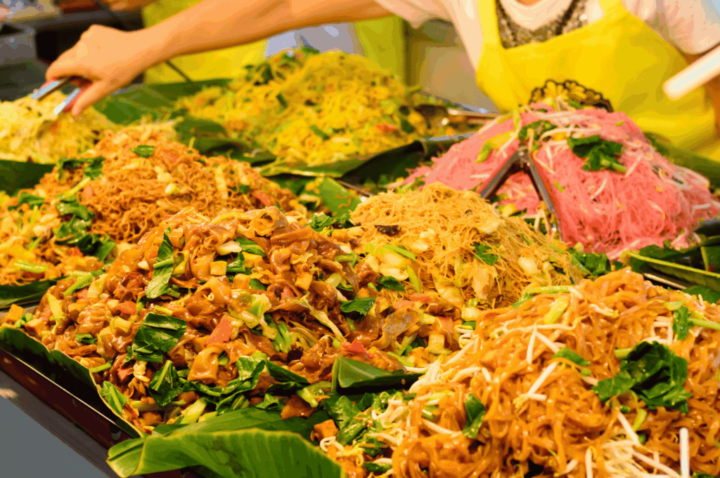 Colorful array of stir-fried Thai noodles displayed on banana leaves at a bustling Bangkok street stall—featuring vibrant yellow, pink, and brown noodles tossed with fresh vegetables and tofu, served by a vendor in a yellow apron, evoking the visual and culinary diversity of Thailand’s must-try street food.