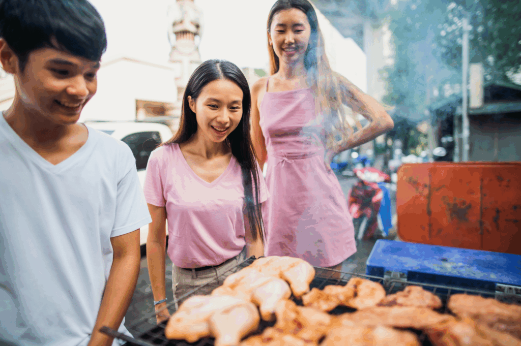 Three friends smiling beside a smoky Bangkok street grill loaded with sizzling chicken skewers—capturing the communal joy, urban grit, and irresistible aroma of Thailand’s open-air food culture.