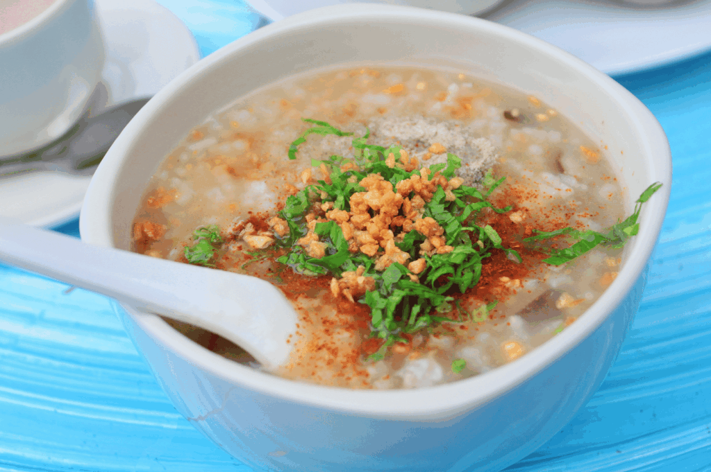 Steaming bowl of Thai rice porridge (jok) garnished with cilantro, fried garlic, black pepper, and chili powder—served with tea on a blue tabletop, evoking the comforting flavors and gentle spice of Bangkok’s beloved breakfast ritual.
