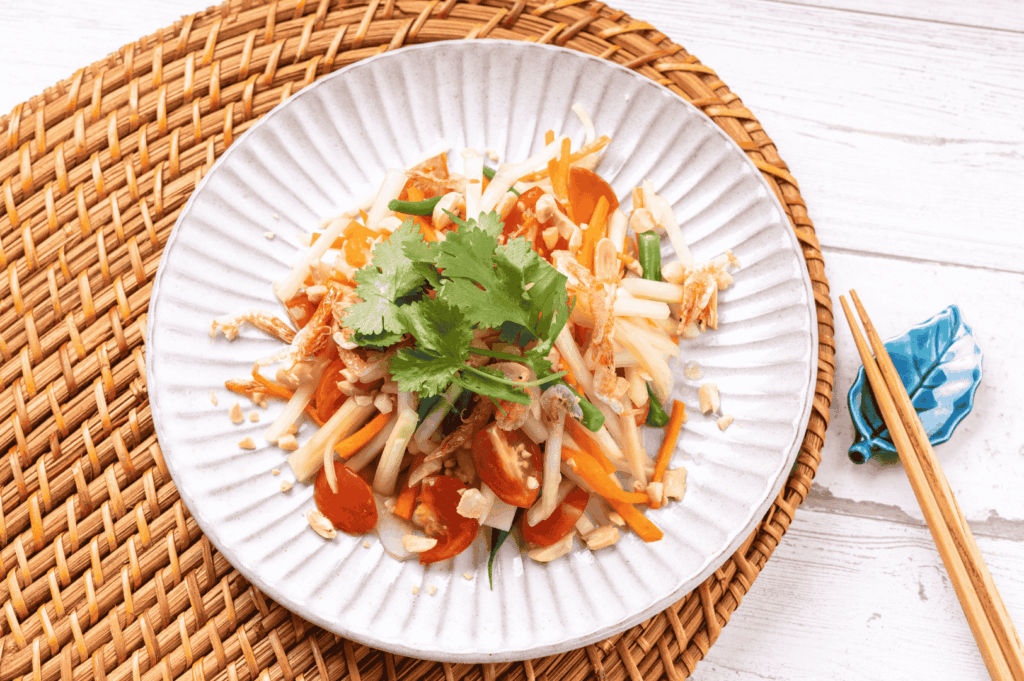 Elegant plate of Thai noodle salad with rice noodles, julienned carrots, red bell peppers, green onions, crushed peanuts, and fresh cilantro—served on a woven mat with wooden chopsticks resting on a blue fish-shaped holder, evoking the freshness and artistry of Bangkok’s lighter street food offerings.