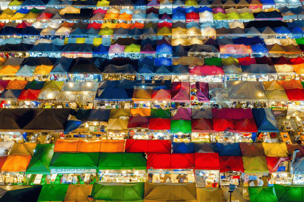 Aerial view of Bangkok’s vibrant night market glowing with rows of multicolored tents—each stall lit from within, forming a kaleidoscopic grid that captures the energy, diversity, and visual spectacle of Thailand’s street food culture after dark.