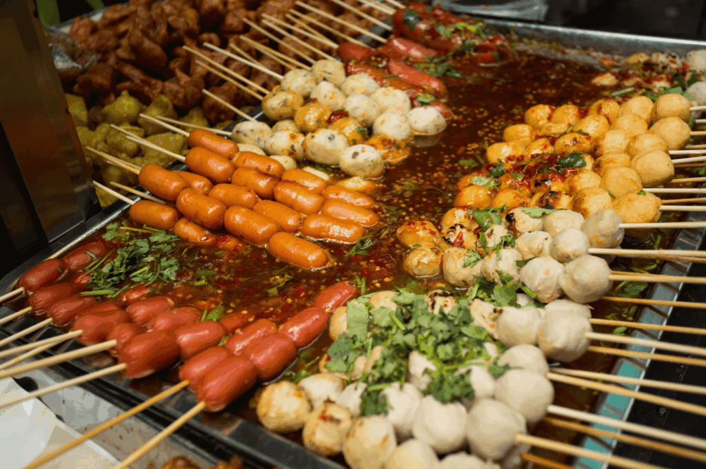 Bangkok street food tray brimming with skewers of sausages, meatballs, and tofu balls soaking in a fiery red chili broth—garnished with cilantro and chili flakes, showcasing the spicy, indulgent edge of Thailand’s street food scene.