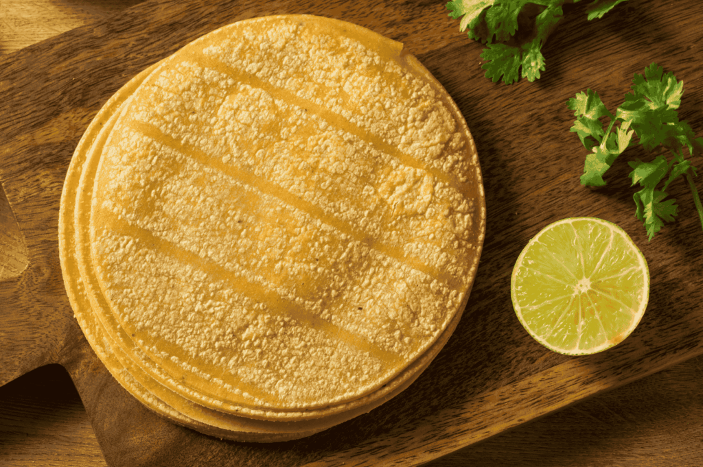Stack of freshly grilled corn tortillas with diagonal char marks, resting on a rustic wooden board beside halved lime and sprigs of cilantro—evoking the warmth and simplicity of homemade Mexican cooking. Ideal visual for illustrating traditional tortilla-making techniques and essential prep elements in a home kitchen setting.