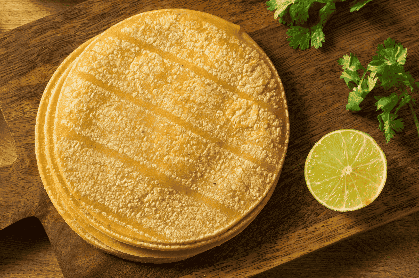 Stack of freshly grilled corn tortillas with diagonal char marks, resting on a rustic wooden board beside halved lime and sprigs of cilantro—evoking the warmth and simplicity of homemade Mexican cooking. Ideal visual for illustrating traditional tortilla-making techniques and essential prep elements in a home kitchen setting.