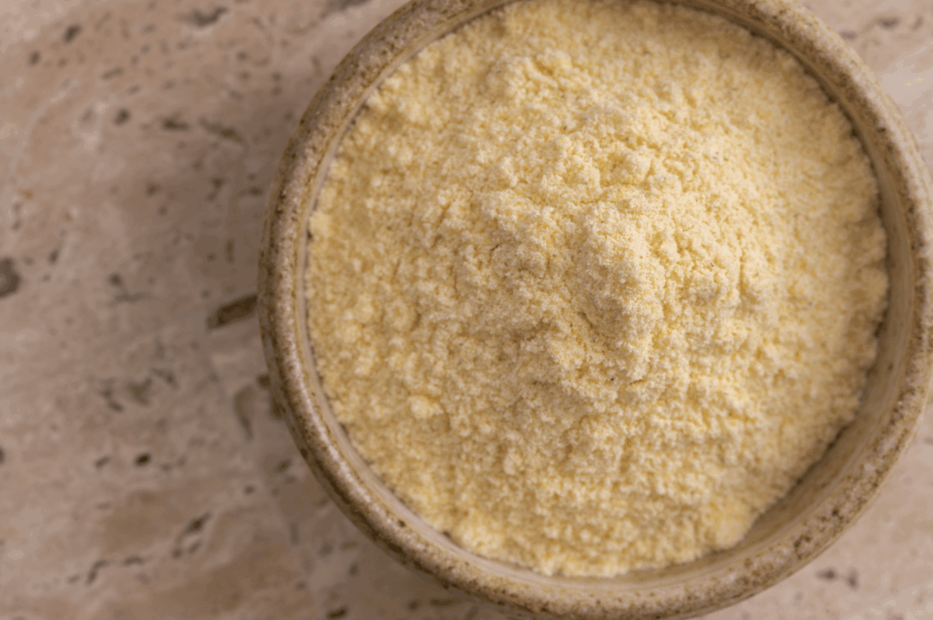 Finely ground yellow cornmeal in a ceramic bowl set on a textured stone surface—highlighting the foundational ingredient for homemade corn tortillas. The soft, granular texture and warm hue evoke the artisanal quality and simplicity of traditional masa preparation.
