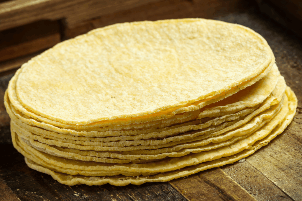 Stack of round, handmade corn tortillas with rustic edges and a slightly rough surface, arranged on a wooden board—capturing the artisanal texture and golden hue of freshly pressed masa. A visual anchor for the tactile satisfaction and tradition behind homemade tortilla preparation.