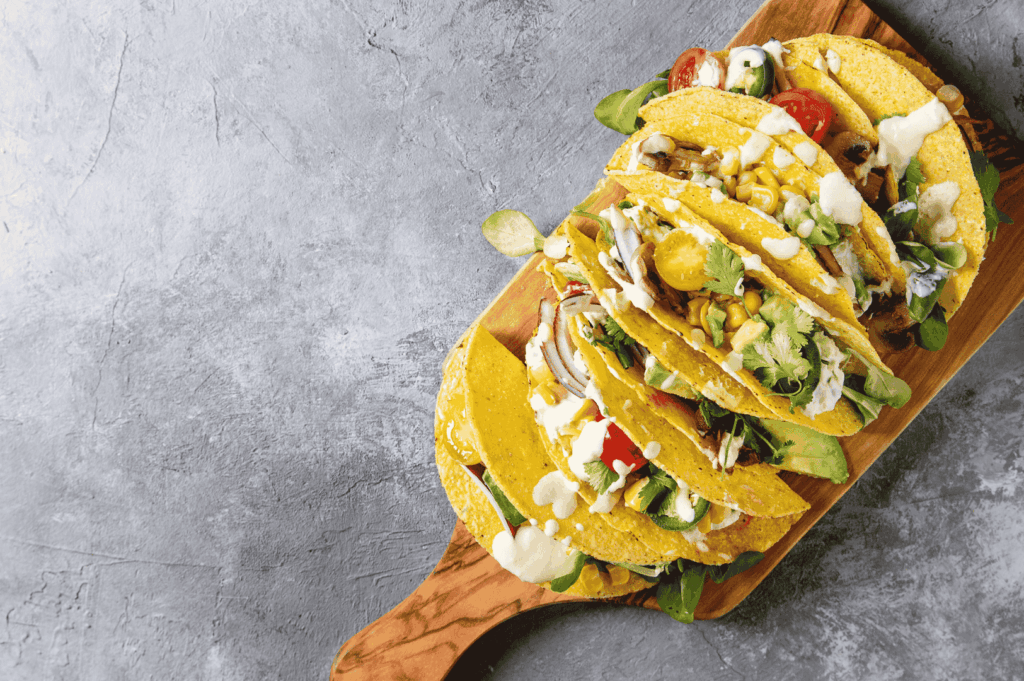 Vibrant vegetarian tacos arranged on a wooden board, each filled with avocado slices, corn kernels, cherry tomatoes, mushrooms, leafy greens, and drizzled with creamy white sauce—wrapped in golden corn tortillas and garnished with fresh herbs. A celebratory finale to the homemade tortilla journey, showcasing the freshness, versatility, and visual appeal of plant-forward Mexican cuisine.
