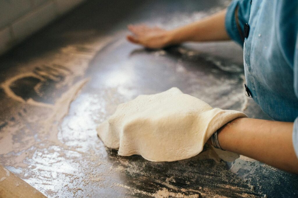 This close-up captures a baker's hands stretching a thin sheet of dough across a flour-dusted metal work surface. Wearing a blue denim-style shirt, the person carefully manipulates the pale dough, emphasizing the tactile and messy nature of the preparation process.