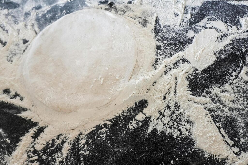 This top-down photograph captures a stack of flattened, circular dough discs resting on a dark surface scattered with loose flour. The high contrast between the pale dough and the black background emphasizes the raw textures and messy preparation process of homemade cooking.