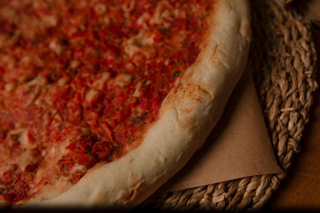 This close-up captures the edge of a rustic pizza, highlighting its thick, pillowy crust and textured red tomato topping sprinkled with herbs. The dish is presented on brown parchment paper over a woven straw placemat, creating a warm and natural aesthetic.