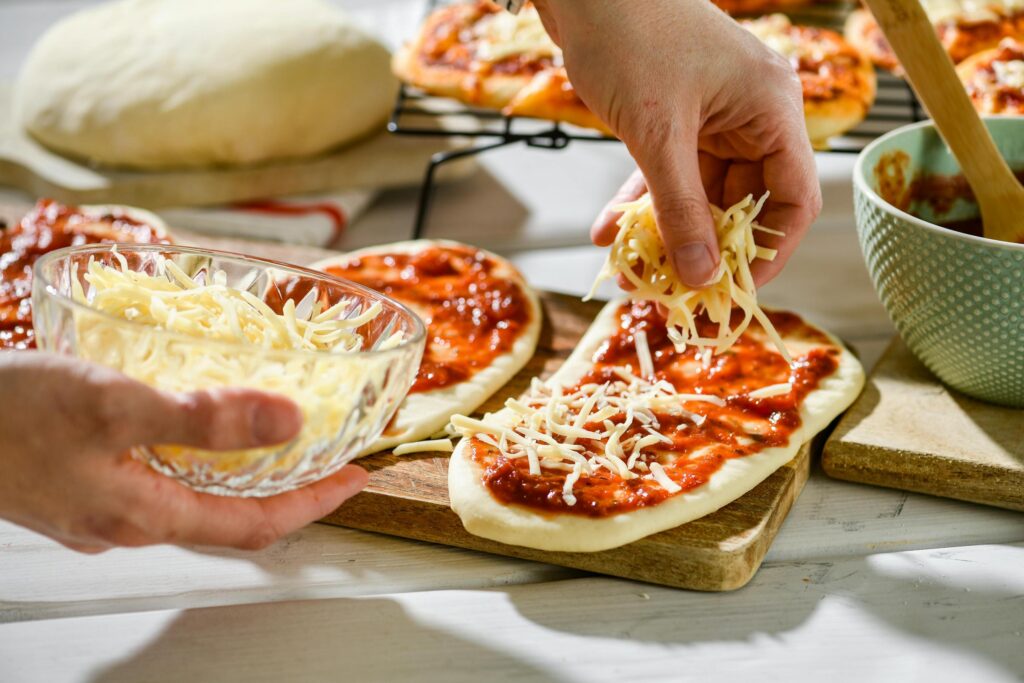 This bright, high-angle shot depicts the preparation of mini pizzas, showing hands sprinkling shredded cheese onto oval dough bases covered in tomato sauce. Surrounding the preparation area on the white wooden table are baking essentials, including a bowl of sauce, a large ball of resting dough, and a rack of finished pastries in the background.