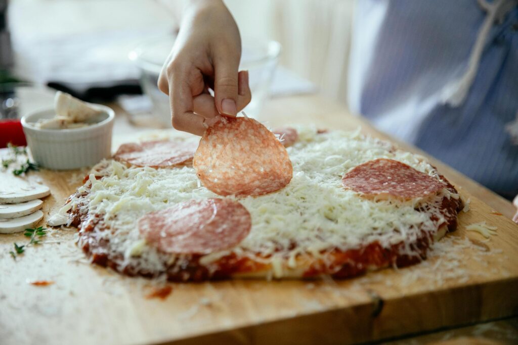 This close-up captures a hand carefully arranging a slice of pepperoni atop a bed of shredded white cheese on a homemade pizza base. The preparation takes place on a wooden board accompanied by small bowls of sliced mushrooms and fresh herbs, evoking a cozy cooking atmosphere.