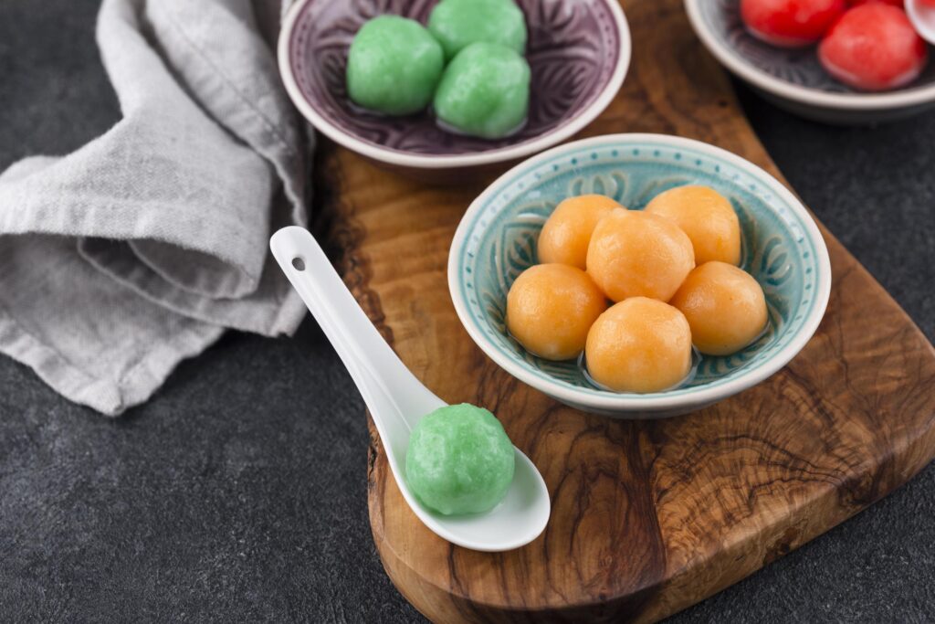 This still life image showcases an assortment of colorful sweet Niangao, featuring a teal bowl filled with orange glutinous rice balls and a purple dish holding green ones. A white ceramic spoon rests on a rustic wooden board displaying a single green ball, sitting beside a gray napkin on a dark surface.