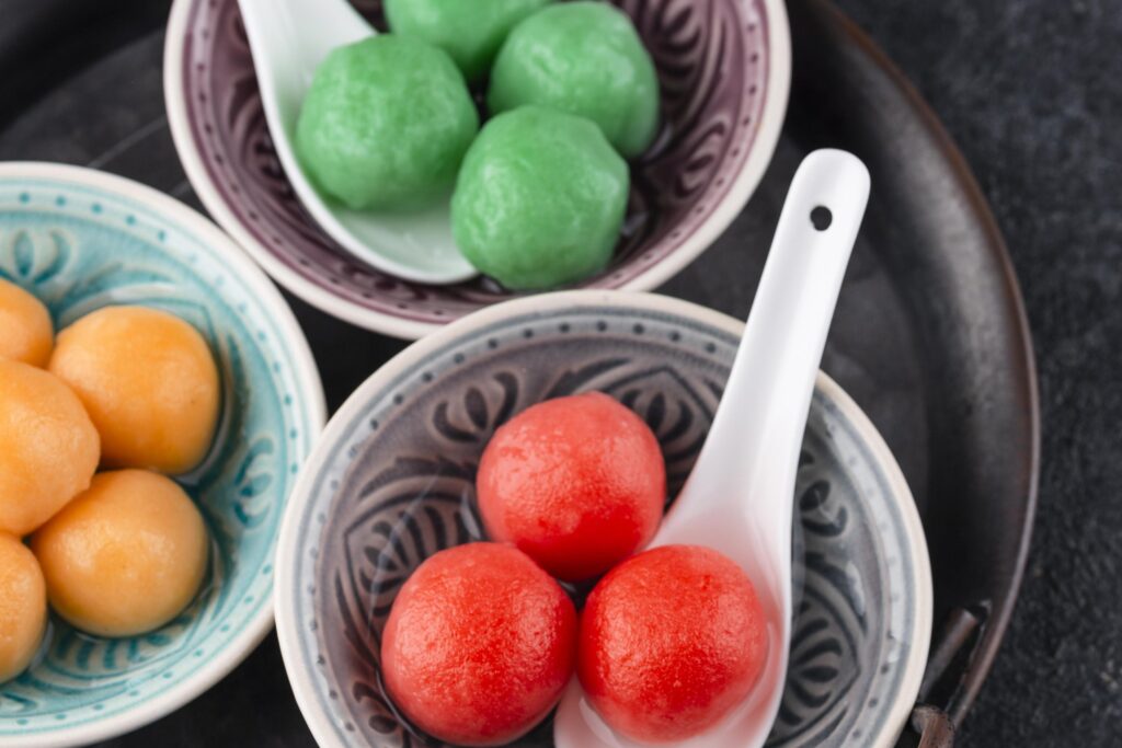This close-up still life captures three patterned bowls arranged on a dark circular tray, containing green, orange, and vibrant red glutinous rice balls. White ceramic spoons rest in the purple and grey dishes, showcasing the glossy texture of the sweet dumplings within the assortment.