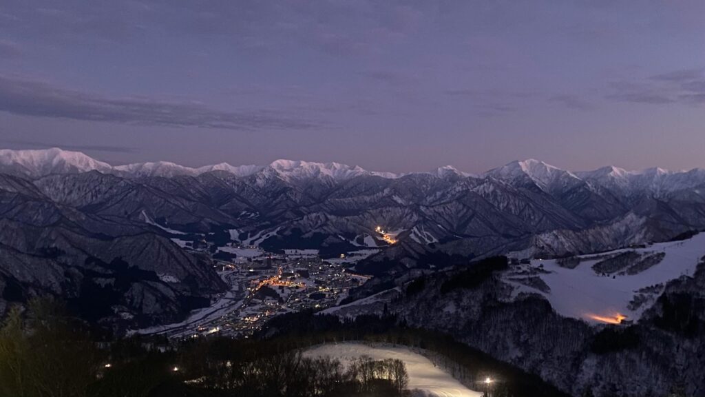 A majestic, snow-covered mountain range stretches across the horizon under a moody, purple twilight sky. Nestled in the dark valley below, a cluster of warm city lights illuminates the town and ski slopes, creating a striking contrast against the cold, rugged landscape.