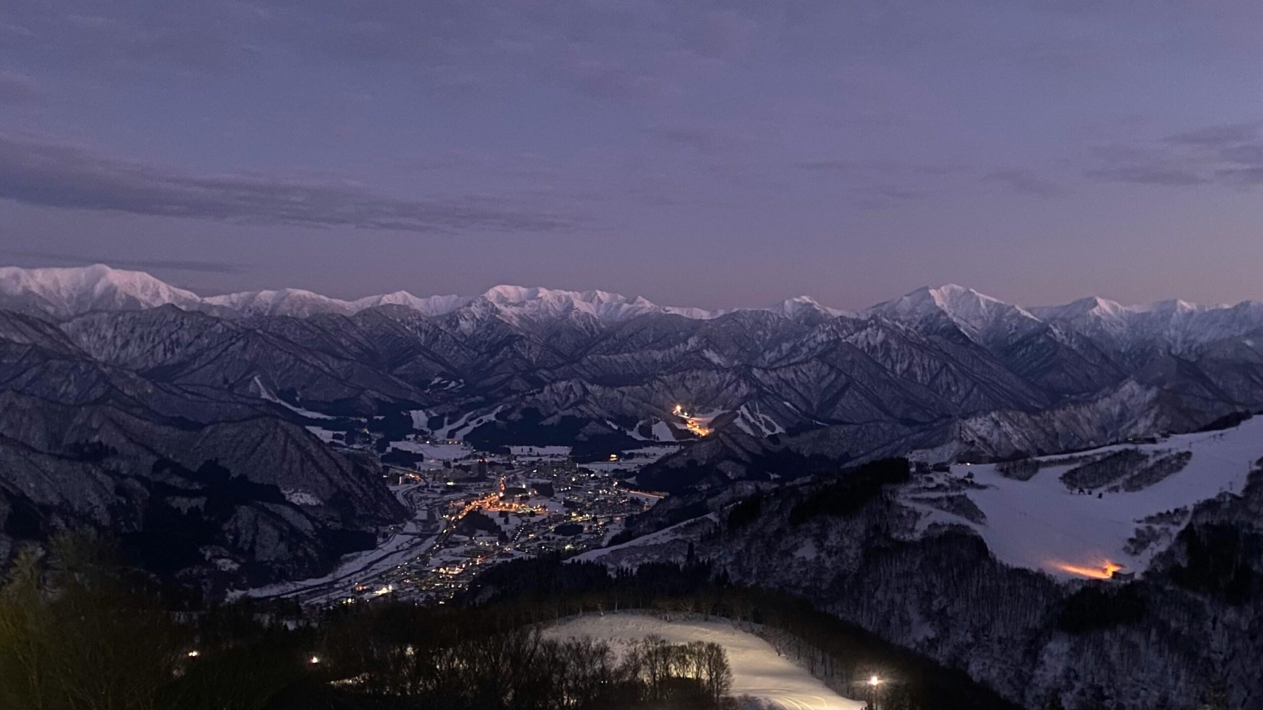 A majestic, snow-covered mountain range stretches across the horizon under a moody, purple twilight sky. Nestled in the dark valley below, a cluster of warm city lights illuminates the town and ski slopes, creating a striking contrast against the cold, rugged landscape.