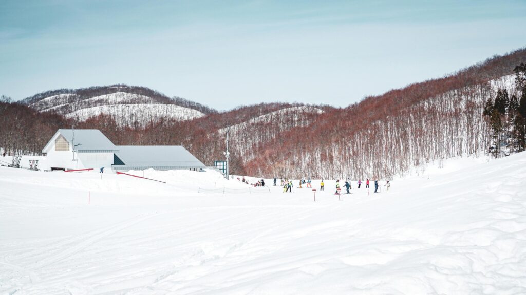 Skiers and snowboarders in colorful winter gear dot a pristine white slope, gathering near a modern lodge facility at the base of a hill. The snowy scene is framed by rolling mountains covered in dense, reddish-brown leafless trees under a pale, bright sky.