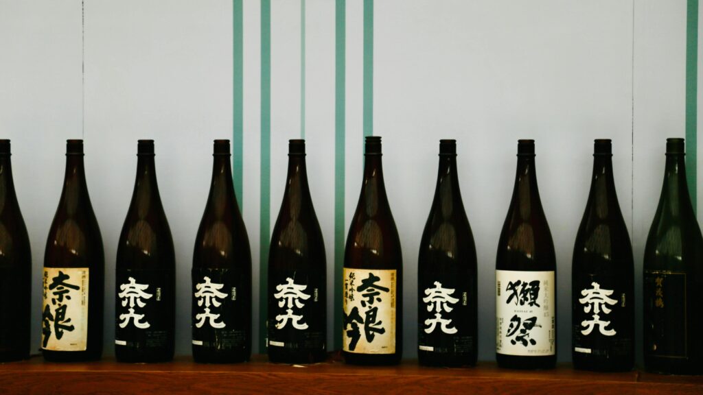 A row of dark glass bottles, likely containing sake, stands neatly on a wooden shelf, displaying labels with bold Japanese calligraphy. The collection is set against a simple background of vertical teal stripes, emphasizing the traditional aesthetic of the packaging.