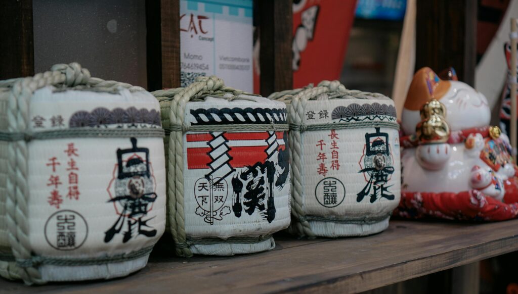 Three traditional sake barrels wrapped in straw rope and adorned with black calligraphy are lined up on a wooden surface. A ceramic lucky cat figurine sits beside them, adding a playful and cultural element to the arrangement.