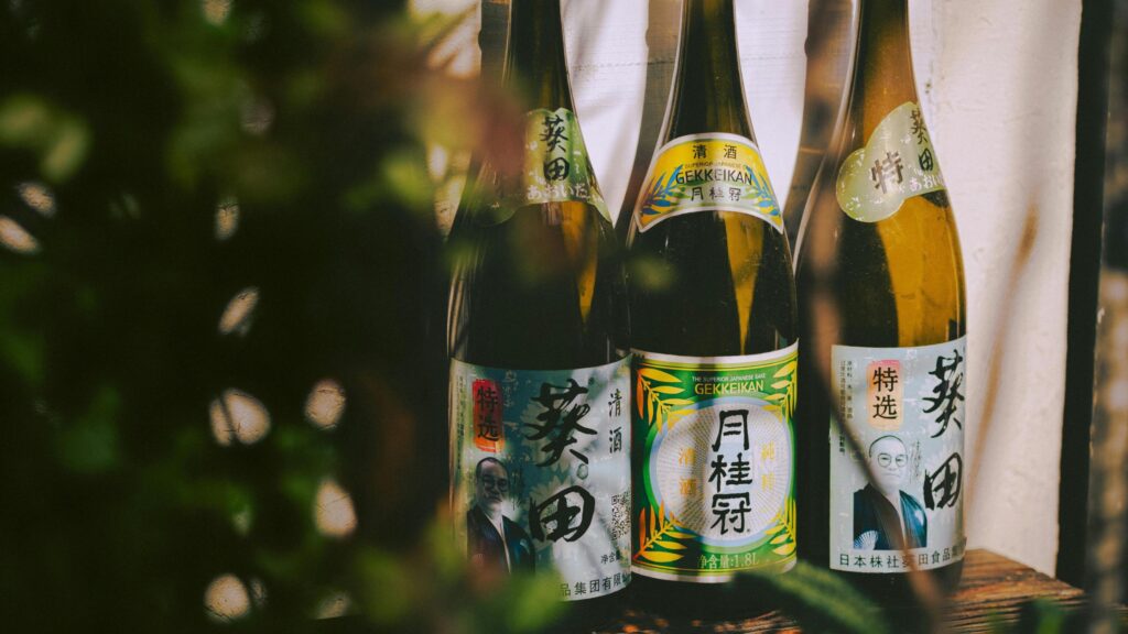 Three large brown glass bottles featuring Japanese calligraphy and distinctive labels, including one for Gekkeikan sake, stand on a wooden surface. Blurred green foliage in the foreground partially obscures the left side of the scene, creating a sense of depth and framing around the traditional beverages.