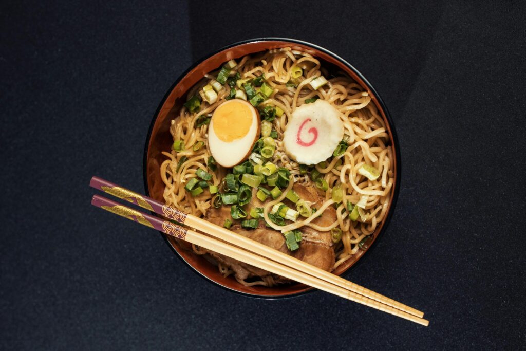 This image features a bowl of ramen topped with sliced duck, two soft-boiled egg halves, and a generous portion of chopped green onions. A black ceramic spoon rests on a matching holder next to the bowl on a light-colored wooden table.