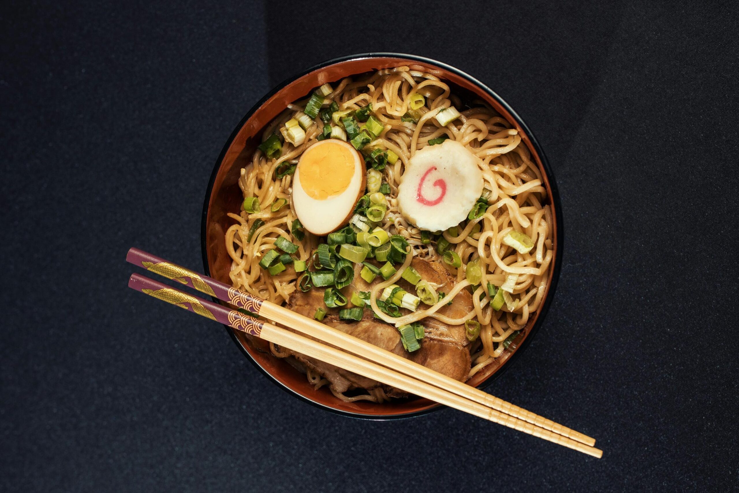 This image features a bowl of ramen topped with sliced duck, two soft-boiled egg halves, and a generous portion of chopped green onions. A black ceramic spoon rests on a matching holder next to the bowl on a light-colored wooden table.
