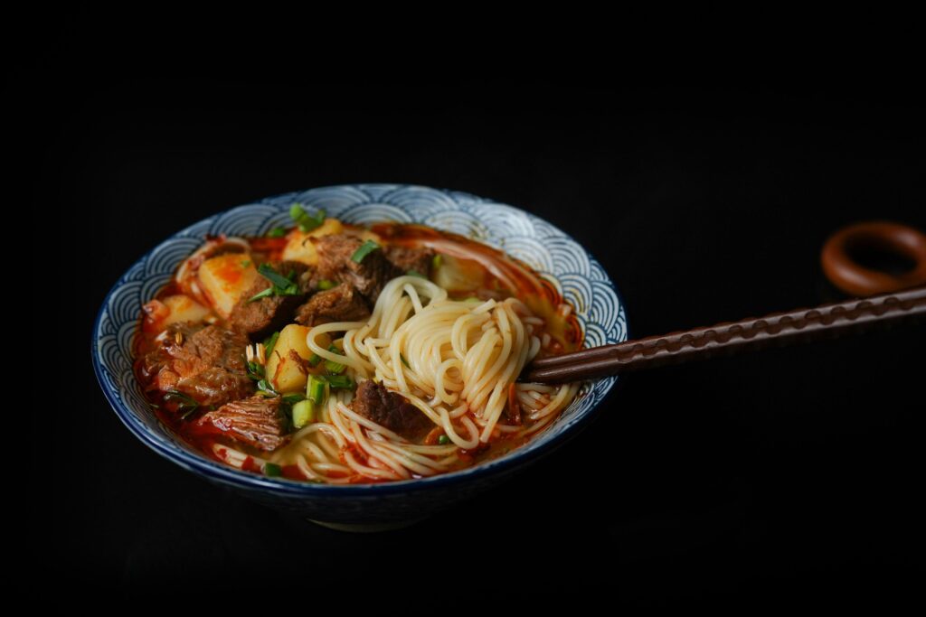 In a patterned blue and white bowl, a spicy red broth is filled with tender white noodles and chunks of braised beef. The dish is garnished with fresh green onions, and a pair of dark, textured chopsticks is shown dipped into the side of the bowl.
