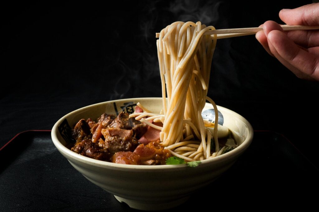 A person's hand uses wooden chopsticks to lift a tall bundle of steaming noodles from a large ceramic bowl filled with dark broth and hearty chunks of meat. The scene is set against a dark background, highlighting the steam rising from the hot dish.