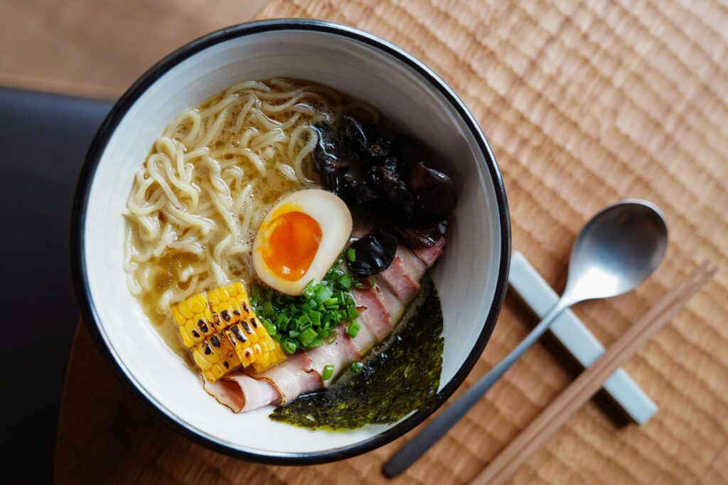 A colorful bowl of ramen features thin, curly noodles topped with a jammy soft-boiled egg, grilled corn, and several slices of pork. The dish is presented on a woven mat and accompanied by a metal spoon and wooden chopsticks resting on a ceramic holder.