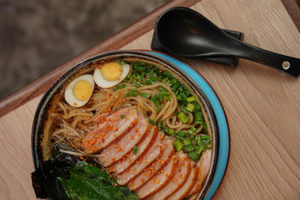 This bowl of ramen is topped with sliced duck seasoned with sesame seeds, two soft-boiled egg halves, and a generous portion of chopped green onions. It is served on a light wooden table alongside a black ceramic soup spoon resting on its matching holder.
