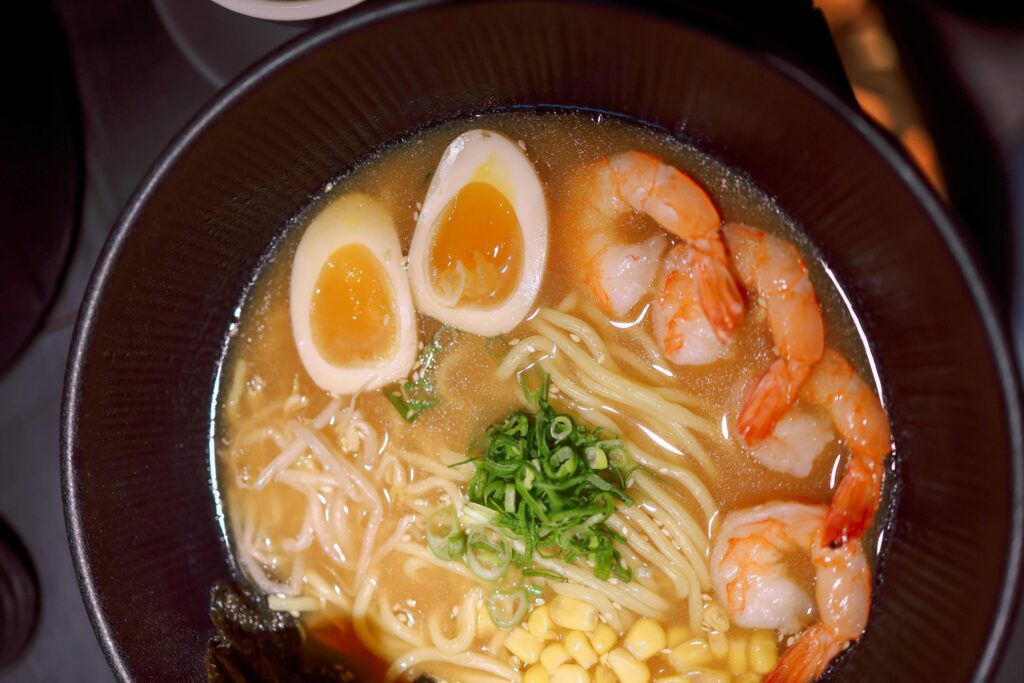 A bowl of shrimp ramen is presented from a top-down perspective, featuring a rich broth filled with noodles and topped with two soft-boiled egg halves. The dish is garnished with several cooked shrimp, corn kernels, bean sprouts, and a sprinkle of fresh green onions.