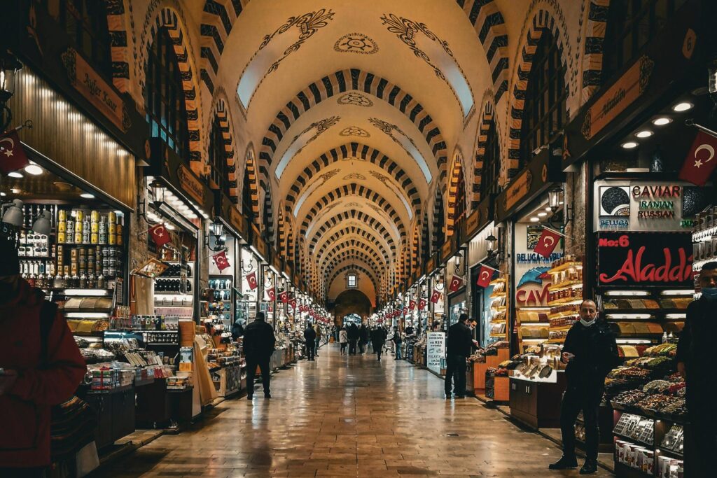 This image depicts the interior of a historic covered bazaar, characterized by soaring arched ceilings with intricate painted designs and alternating light and dark stonework. The spacious, polished corridor is lined with brightly lit shops selling spices, sweets, and souvenirs, adorned with Turkish flags hanging prominently along the walkway.