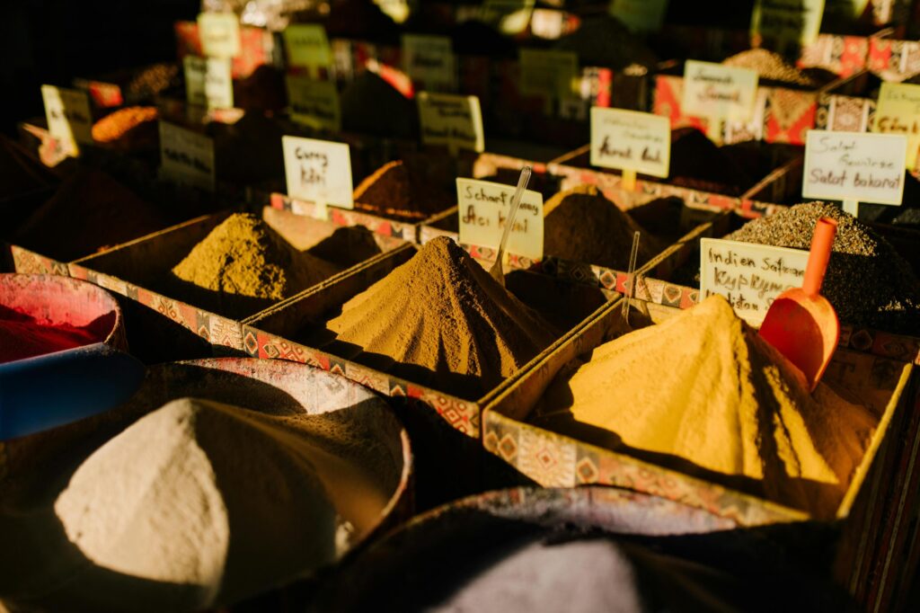 This image features a market stall display with rows of square bins containing mounds of powdered spices, illuminated by strong sunlight that creates distinct highlights and shadows. Small yellow handwritten labels identify the various seasonings, such as curry and saffron, while plastic scoops rest within the heaps.