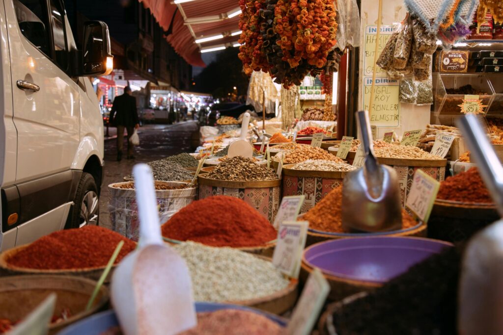 This image captures a street-side market stall overflowing with open baskets of vibrant spices, seeds, and nuts, complete with metal scoops for serving. To the left, a white van is parked next to the display, while strings of dried goods hang above the stall facing a dimly lit street.