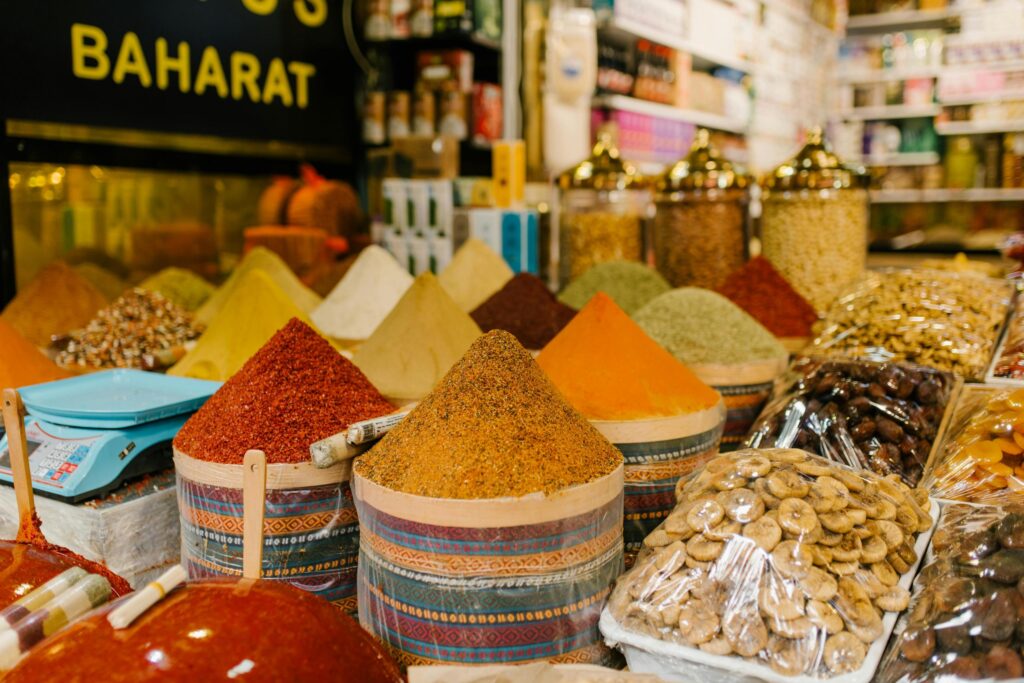 This close-up image showcases a vibrant market display featuring large, cone-shaped mounds of colorful spices, such as red pepper and curry powder, sitting in patterned containers. Adjacent to the spices are trays of dried figs and other fruits wrapped in plastic, with a blue digital scale and shelves of glass jars visible in the blurred background.