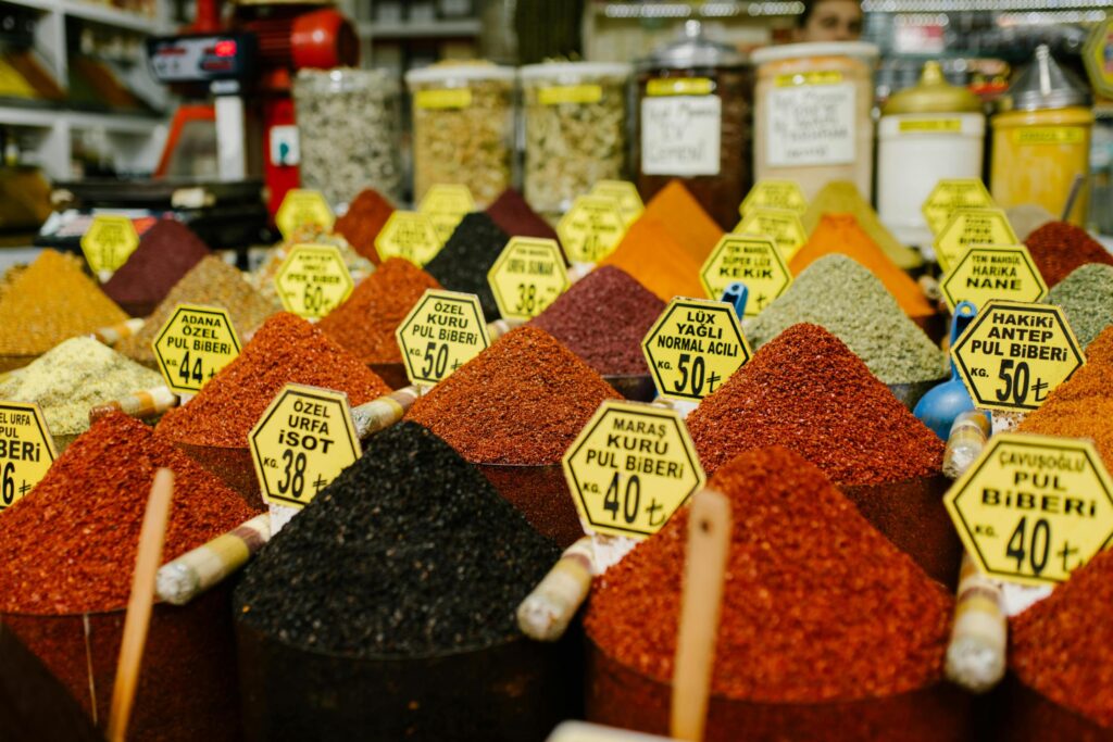 This close-up shot highlights a densely packed display of colorful spice mounds, primarily in shades of red and black, shaped into perfect cones. Bright yellow hexagonal price tags with Turkish text protrude from the peaks of the spices, identifying different varieties of peppers and seasonings amidst the crowded stall.