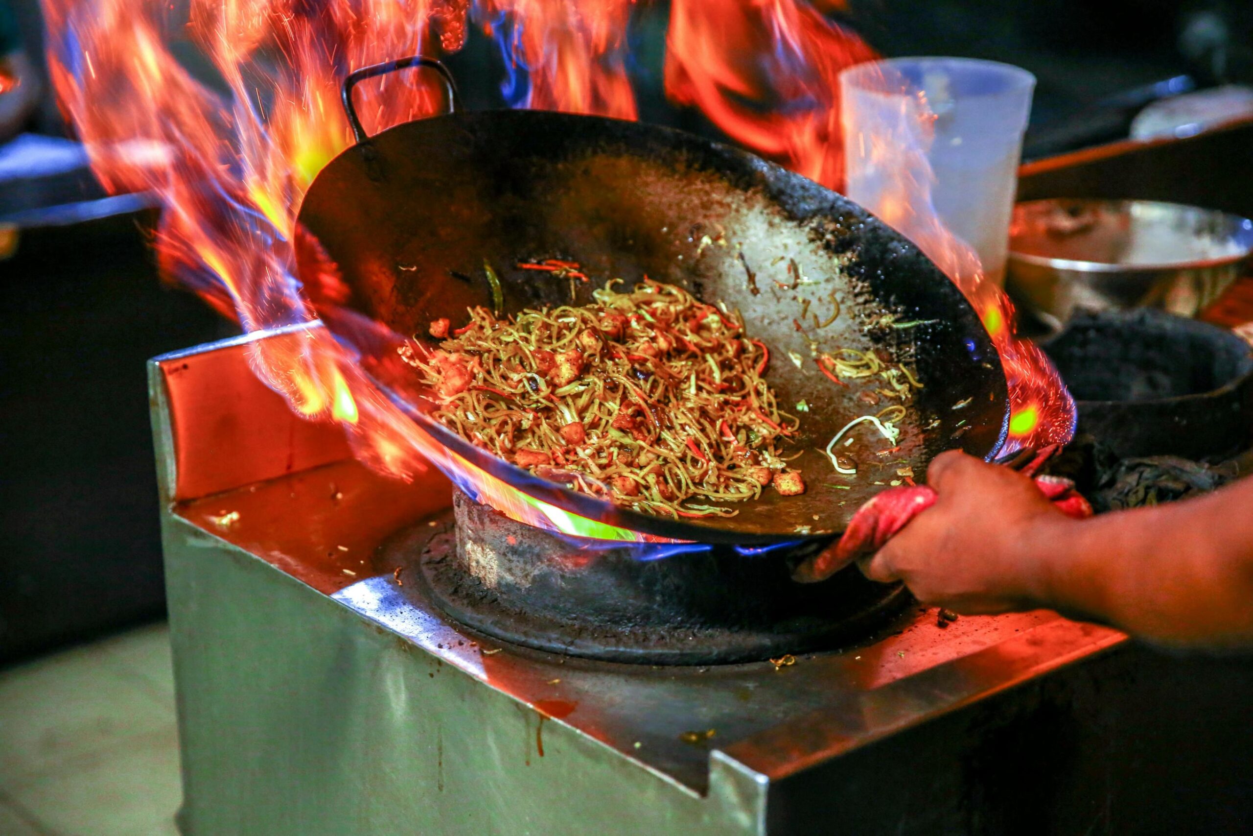 A large wok filled with noodles and vegetables is engulfed in dramatic flames as a cook actively tosses the food over a high-heat burner. The scene captures the intense energy and traditional technique of stir-frying in a professional kitchen setting.