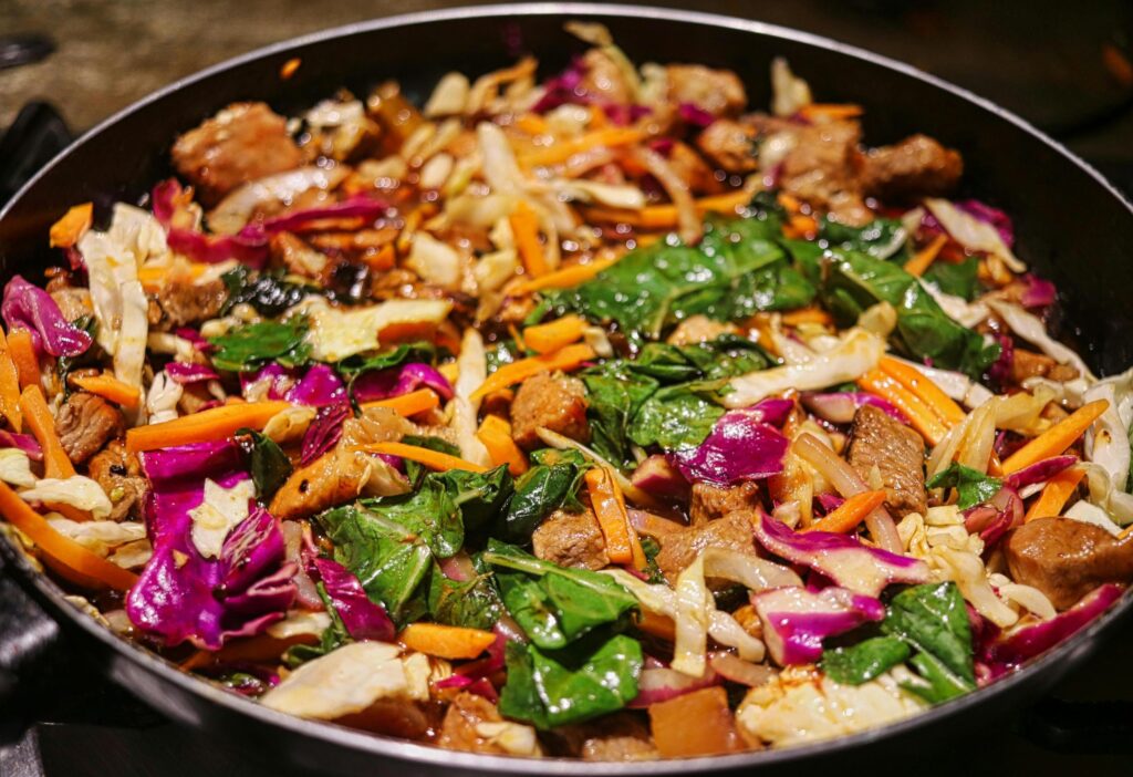 A vibrant stir-fry cooks in a large pan, combining chunks of browned meat with a colorful array of red cabbage, shredded carrots, and green leafy vegetables. The glistening ingredients appear evenly coated in sauce, showcasing a hearty and textural mix of fresh produce.