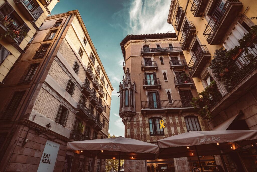 Captured from a dramatic low angle, historic European buildings with warm stone facades and ornate iron balconies tower against a bright blue sky. At street level, large beige umbrellas shelter an outdoor tapas bar, anchoring the architectural grandeur with a cozy, inviting atmosphere.