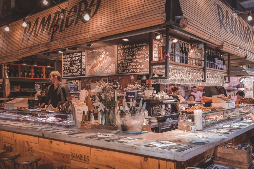 Beneath bamboo shades stamped with "Ramblero," a busy seafood stall displays a chalkboard menu of fresh catches like grilled lobster and sea bass. The lively scene features staff working behind a counter filled with seafood on ice, surrounded by hanging wine glasses and rustic wooden decor.