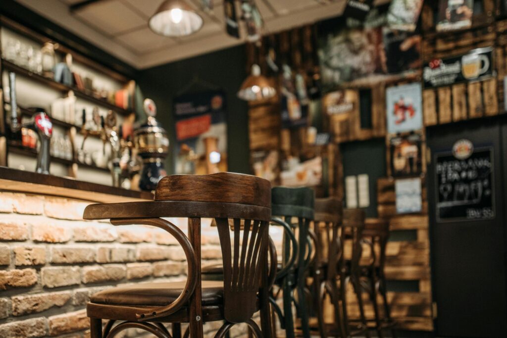 Classic wooden bar stools with curved backs line a textured brick counter, standing in sharp focus within a dimly lit pub. The rustic background features blurred beer taps and vintage wall decorations, creating a warm and inviting atmosphere.
